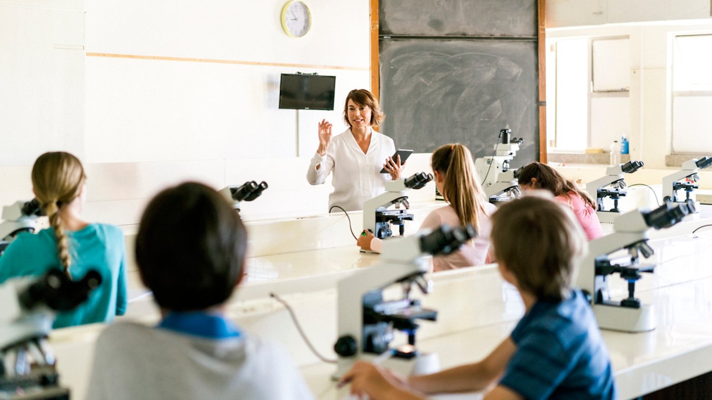 A photo of teacher explaining students by using digital tablet. Boys and girls are sitting with microscope at desk. They are in science class at school laboratory.