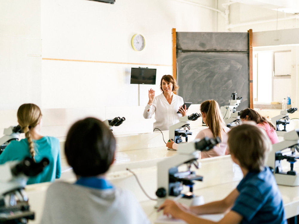 A photo of teacher explaining students by using digital tablet. Boys and girls are sitting with microscope at desk. They are in science class at school laboratory.