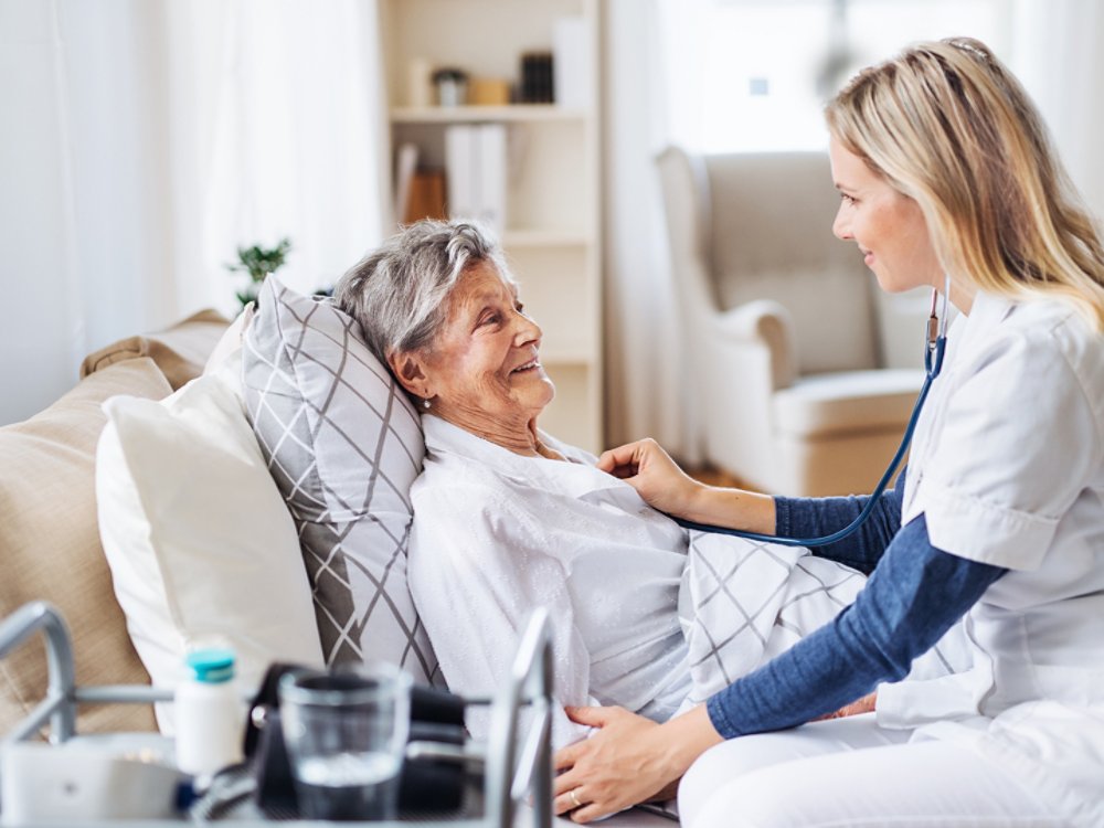 A young health visitor examining a sick senior woman lying in bed at home with stethoscope.