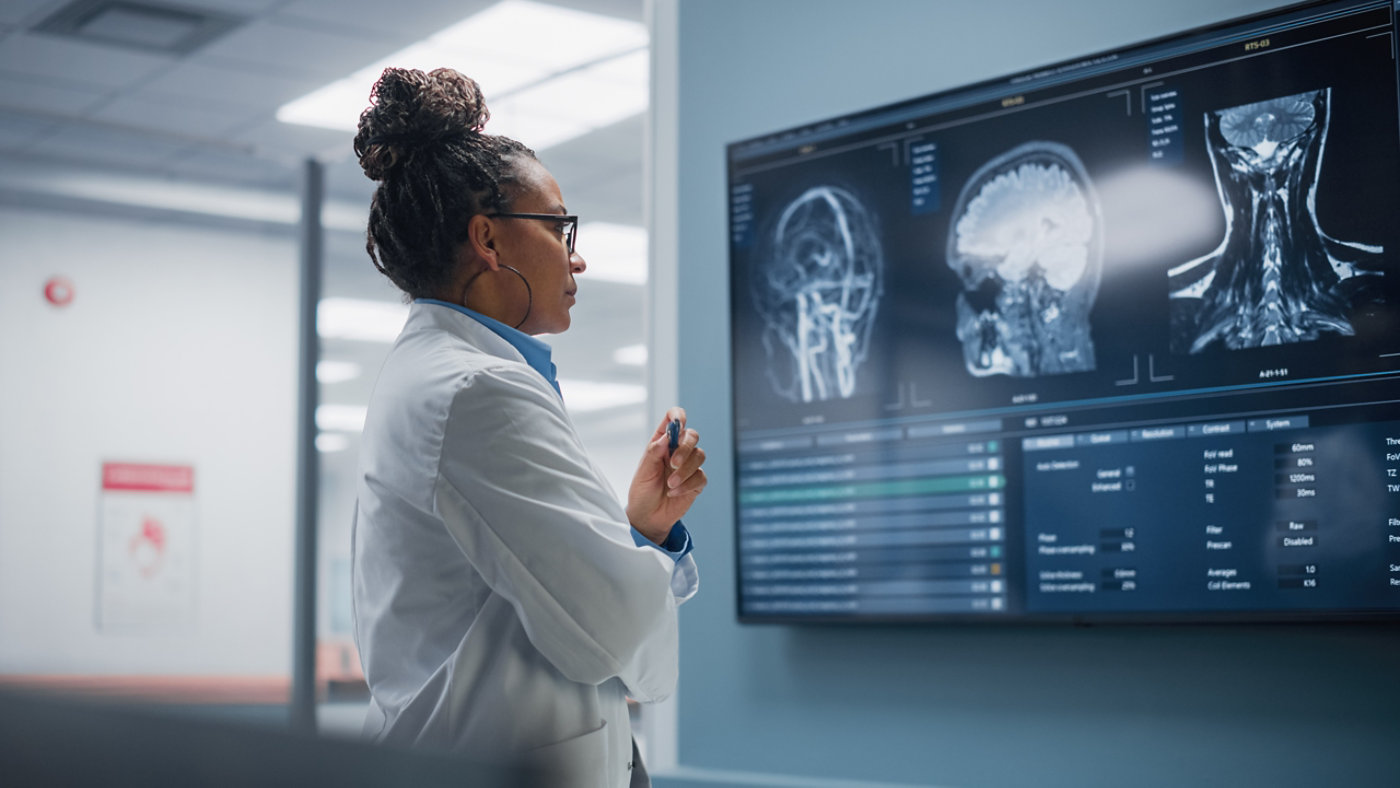 A medical professional in a white lab coat examines detailed MRI images of a brain and neck displayed on a large digital screen. The setting appears to be a modern hospital or diagnostic imaging room, with clinical lighting and advanced technology. The scene conveys a sense of focus and analysis, with the doctor standing attentively in front of the medical images. No visible text or numbers are clearly identifiable on the screen.