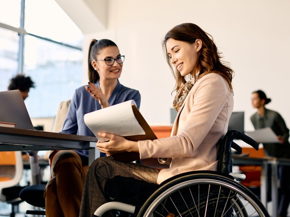 Happy businesswoman in wheelchair going through reports while working female coworker in the office. 