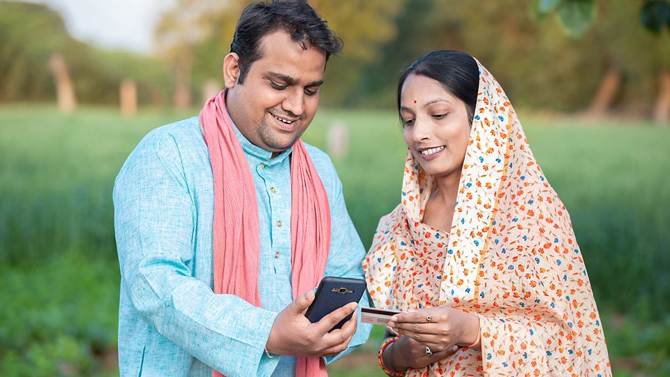 Happy indian rural farmer couple using smartphone to make online payment with debit card in agricultural field, shopping on internet with cellphone secure banking service system concept.