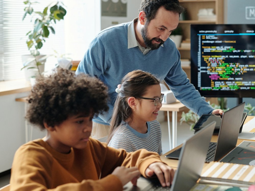 Teacher instructing a diverse group of children coding on laptops in classroom setting. Children eagerly engaging with programming on laptops