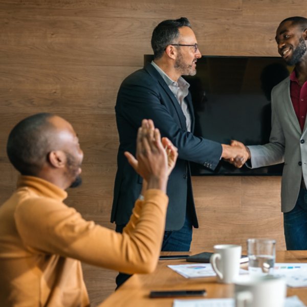 Two diverse businessmen shaking hands while coworkers clap in congratulations