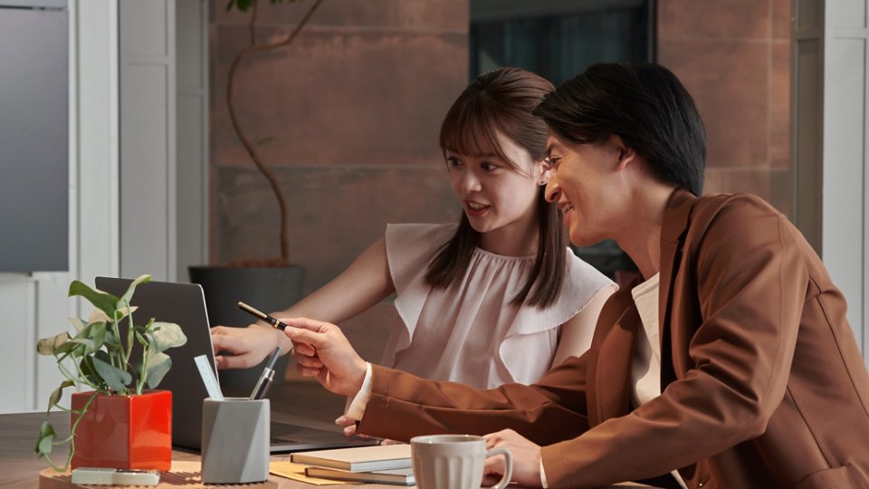 Two colleagues sit together at a wooden desk, discussing something on a laptop screen. One person points at the laptop while the other gestures with a pen, suggesting active collaboration. A potted plant, notebooks, and a coffee mug add a casual, contemporary feel to the workspace. Warm indoor lighting and a neutral background create a professional yet relaxed office atmosphere.