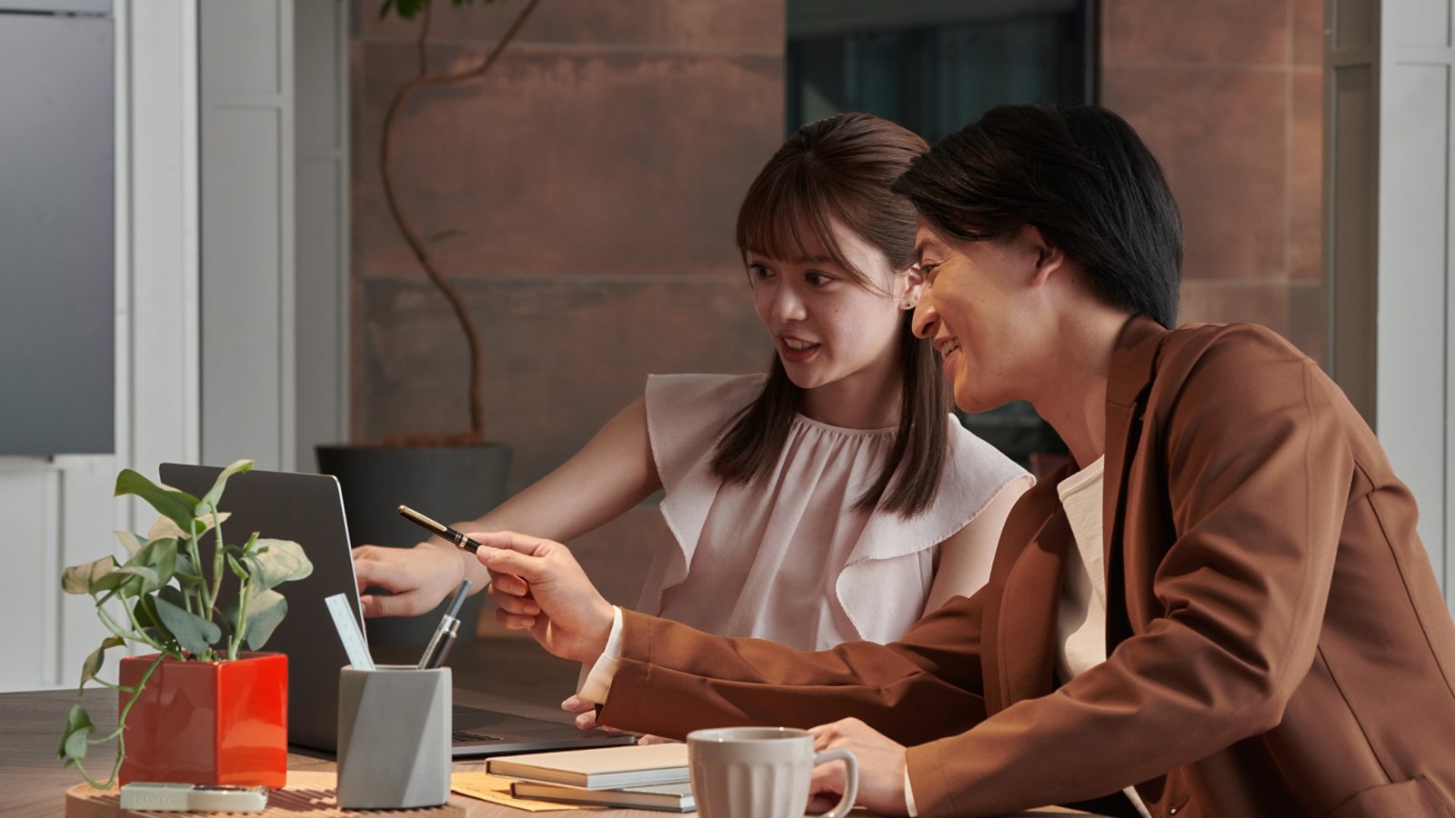 Two colleagues sit together at a wooden desk, discussing something on a laptop screen. One person points at the laptop while the other gestures with a pen, suggesting active collaboration. A potted plant, notebooks, and a coffee mug add a casual, contemporary feel to the workspace. Warm indoor lighting and a neutral background create a professional yet relaxed office atmosphere.