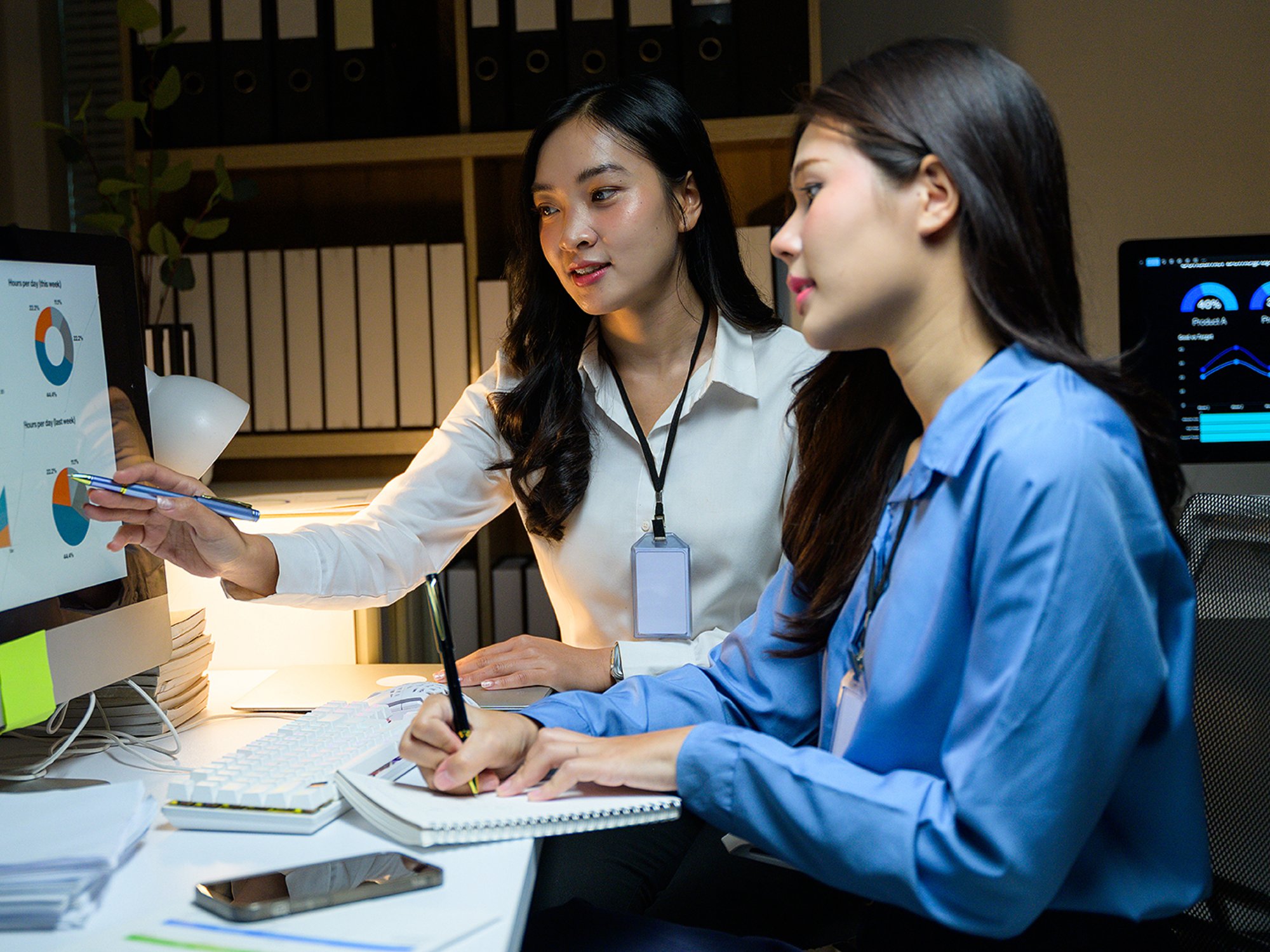 Two Asian businesswomen analyzing financial data on a computer screen, working late in the office