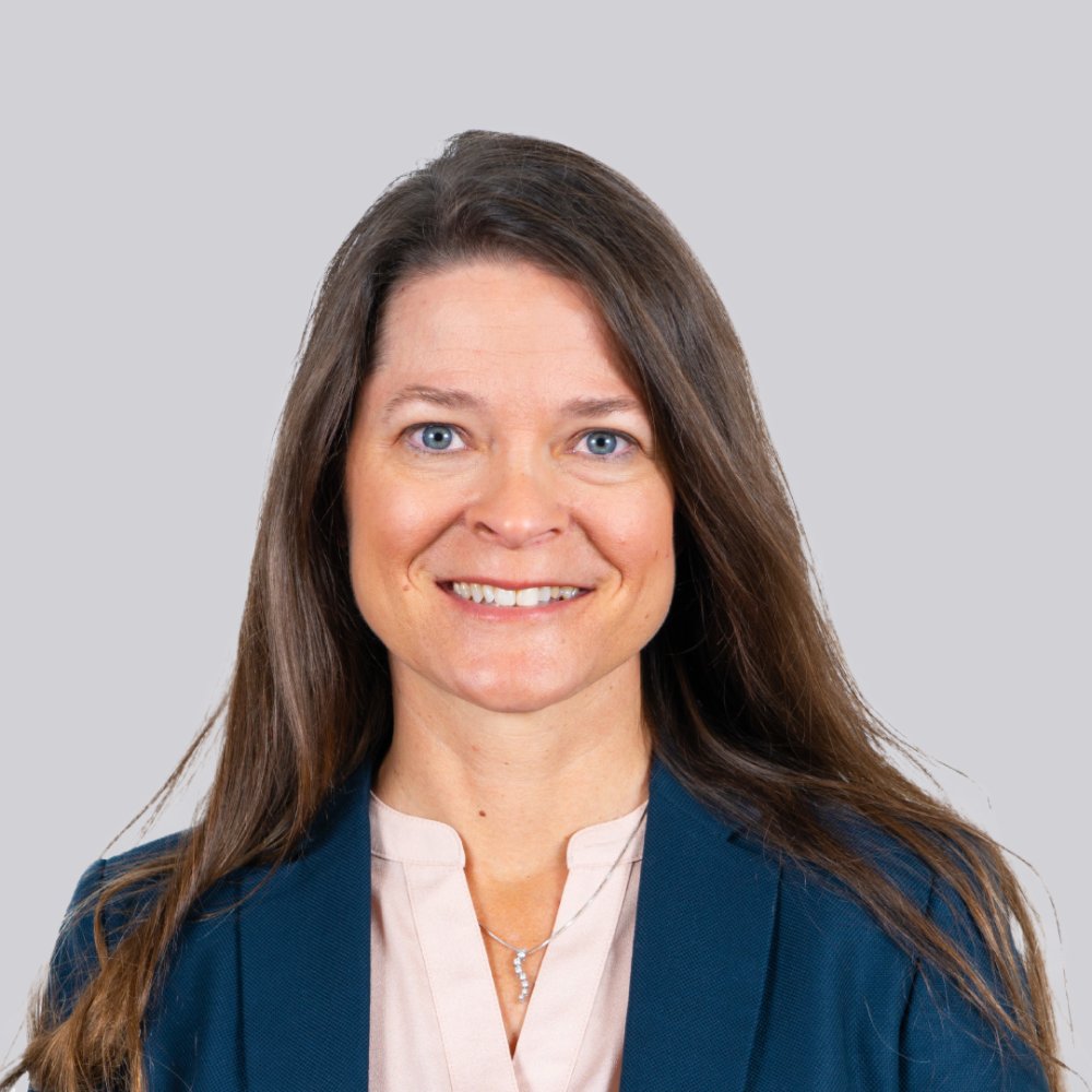 A woman with long brown hair is dressed in a navy blazer and light pink blouse. She is positioned against a neutral gray background, creating a clean and professional look. The outfit suggests a formal or corporate setting.