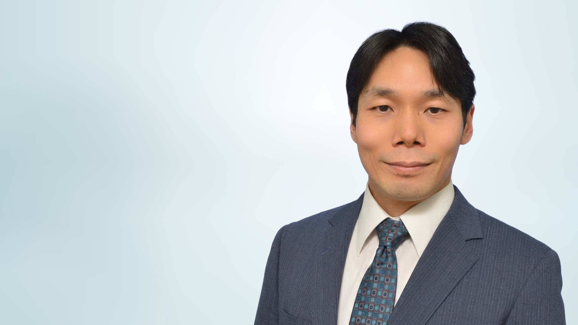 Formal studio portrait of a businessman wearing a dark blue suit, white shirt, and patterned tie. He is facing the camera with a neutral, confident expression. The background is a soft, light gradient that keeps the focus on the subject. The image conveys professionalism and corporate style suitable for business profiles or presentations.