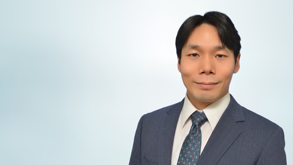 Formal studio portrait of a businessman wearing a dark blue suit, white shirt, and patterned tie. He is facing the camera with a neutral, confident expression. The background is a soft, light gradient that keeps the focus on the subject. The image conveys professionalism and corporate style suitable for business profiles or presentations.
