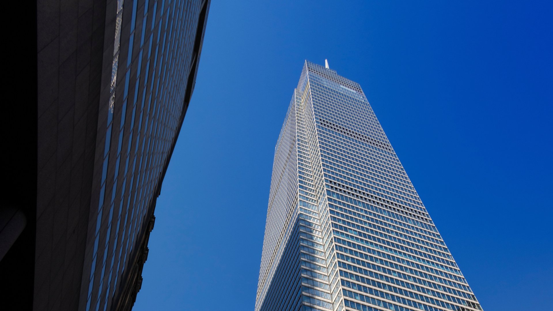 A towering modern skyscraper with a sleek glass facade rises against a vibrant blue sky. The perspective emphasizes its height and architectural design, with another building partially visible in the foreground. The image captures urban sophistication and contemporary style.