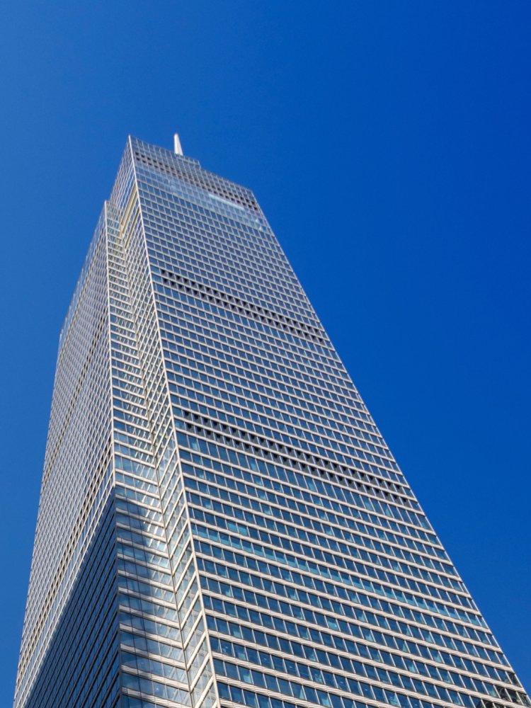 A towering modern skyscraper with a sleek glass facade rises against a vibrant blue sky. The perspective emphasizes its height and architectural design, with another building partially visible in the foreground. The image captures urban sophistication and contemporary style.