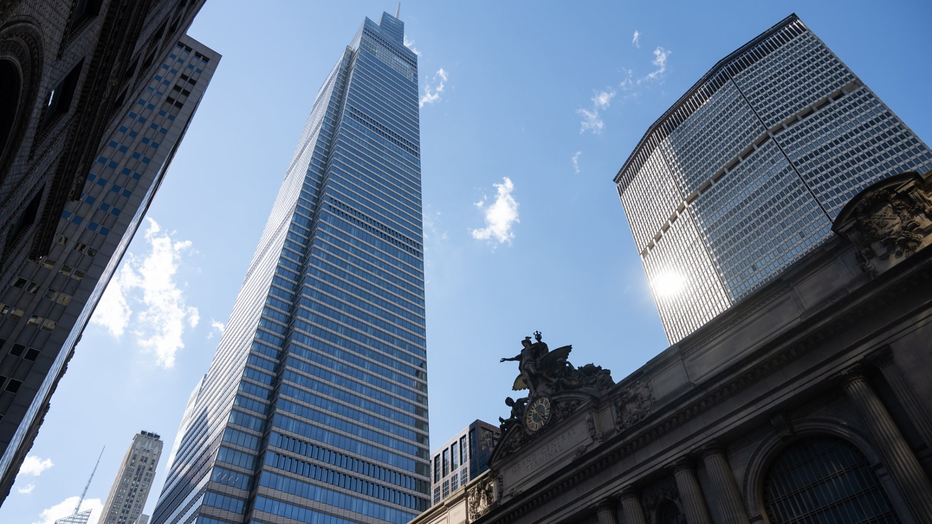 A sleek glass skyscraper rises prominently against a bright blue sky with scattered clouds. The scene is set in a dense urban environment, framed by neighboring high-rise office buildings. In the foreground, an ornate historic structure with sculptural details contrasts with the modern architecture. Sunlight reflects off the glass surfaces, emphasizing the vertical lines and contemporary cityscape. The overall mood is dynamic and metropolitan, highlighting the blend of old and new architecture.