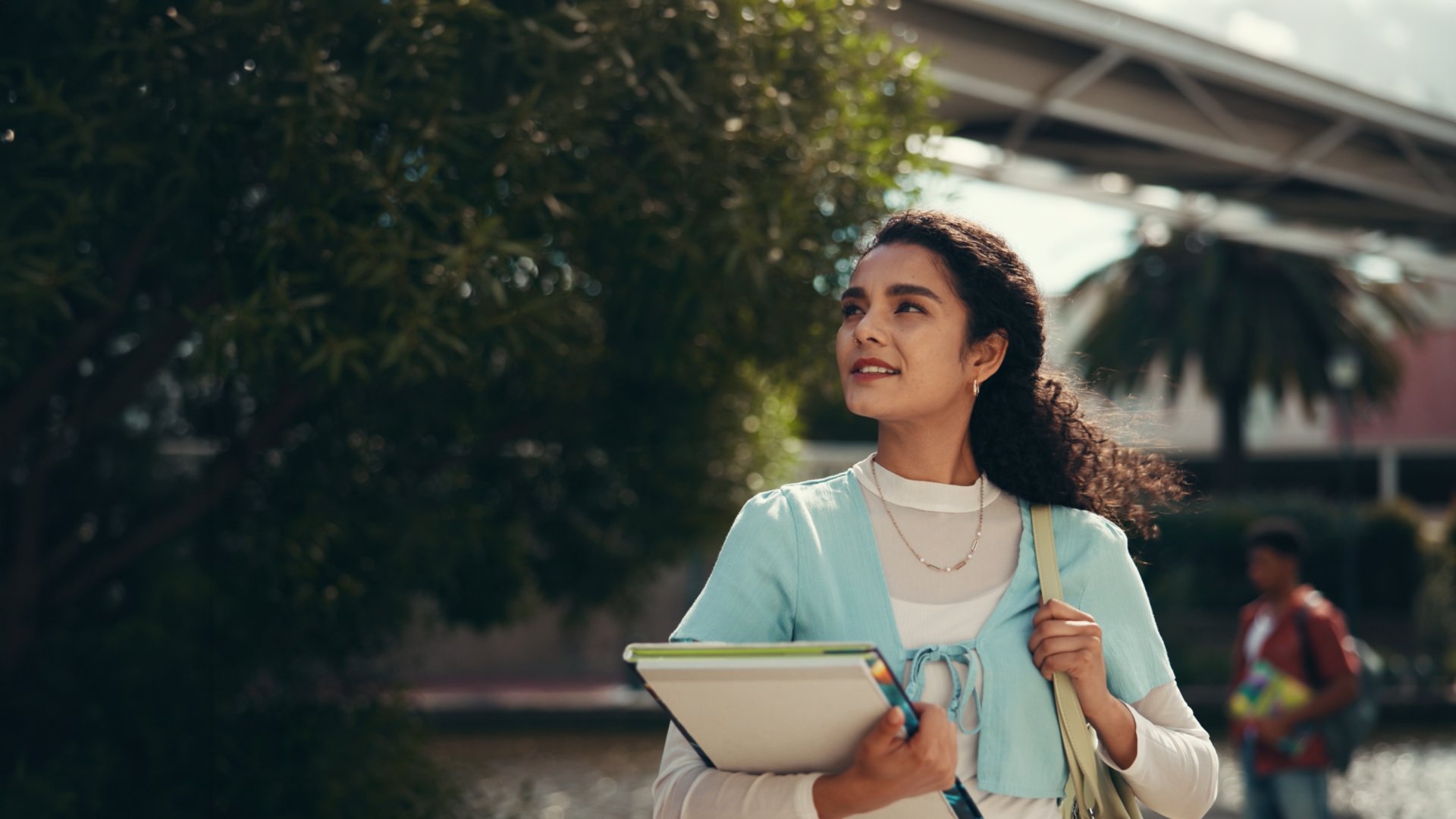 A young woman walks outdoors on a campus, holding notebooks and folders against her chest. She looks upward with a confident expression, surrounded by trees and modern buildings. The background includes another student and architectural structures, suggesting an academic environment. Warm sunlight and soft colors create an optimistic, aspirational mood.