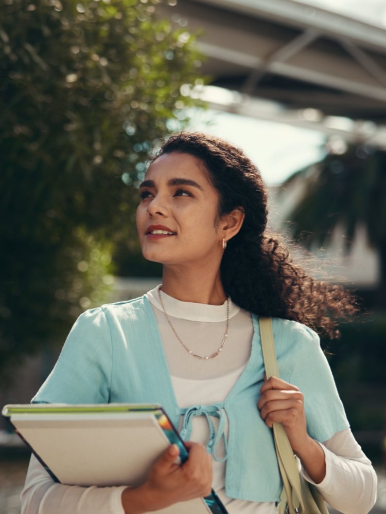 A young woman walks outdoors on a campus, holding notebooks and folders against her chest. She looks upward with a confident expression, surrounded by trees and modern buildings. The background includes another student and architectural structures, suggesting an academic environment. Warm sunlight and soft colors create an optimistic, aspirational mood.