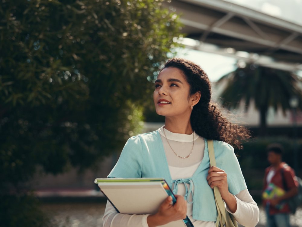 A young woman walks outdoors on a campus, holding notebooks and folders against her chest. She looks upward with a confident expression, surrounded by trees and modern buildings. The background includes another student and architectural structures, suggesting an academic environment. Warm sunlight and soft colors create an optimistic, aspirational mood.
