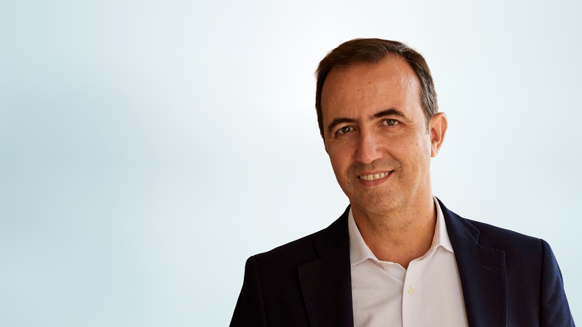 Portrait of a middle-aged man wearing a dark blazer and light-colored shirt. He is smiling gently and looking at the camera, suggesting a professional yet approachable demeanor. The background is a soft, light gradient that keeps the focus on the subject and conveys a clean, corporate style. The overall image feels modern and suitable for business or executive profiles.