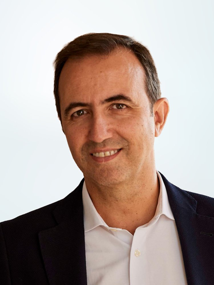 Portrait of a middle-aged man wearing a dark blazer and light-colored shirt. He is smiling gently and looking at the camera, suggesting a professional yet approachable demeanor. The background is a soft, light gradient that keeps the focus on the subject and conveys a clean, corporate style. The overall image feels modern and suitable for business or executive profiles.