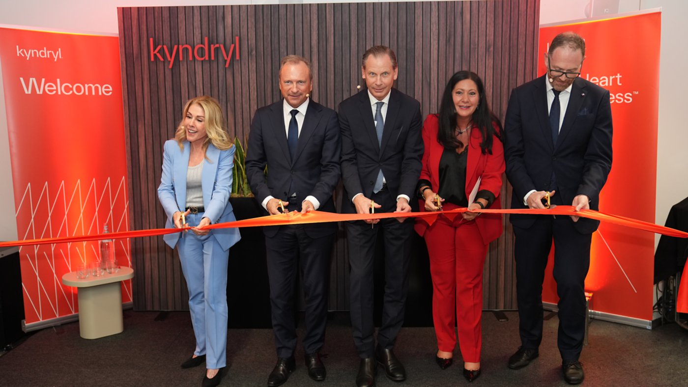 A group of five business professionals stand in a row cutting a ceremonial red ribbon. They are dressed in formal business attire, with two women and three men participating in the ribbon-cutting. The backdrop features a wood-textured wall with illuminated orange panels on each side. The word "kyndryl" appears prominently on the wall, and the word "Welcome" is visible on the left panel. The scene suggests a corporate opening or launch celebration in a modern indoor setting.