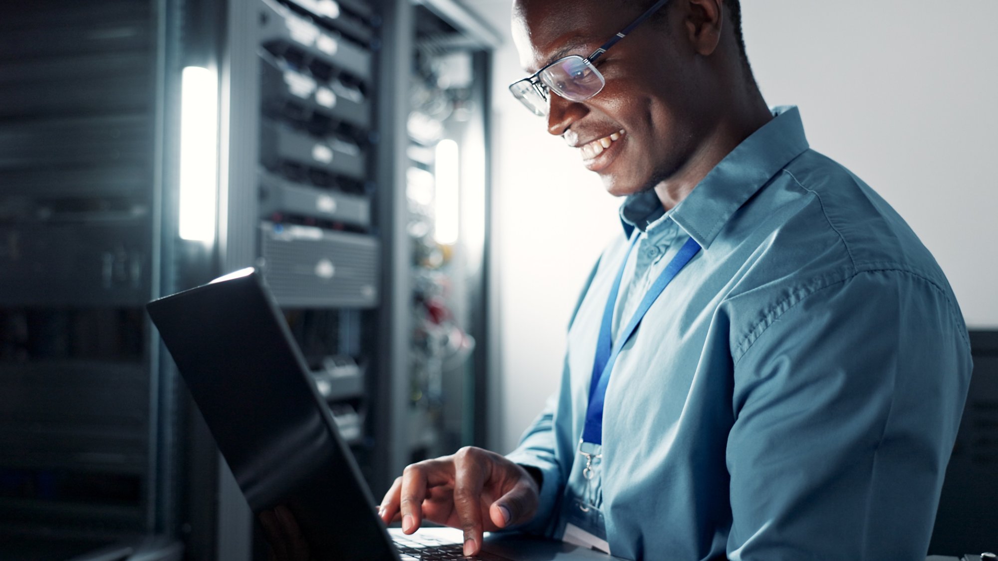 Laptop, server room and black man on database network for maintenance, programming software or typing. Tech, data center and happy engineer on system for information, cybersecurity or cloud computing.