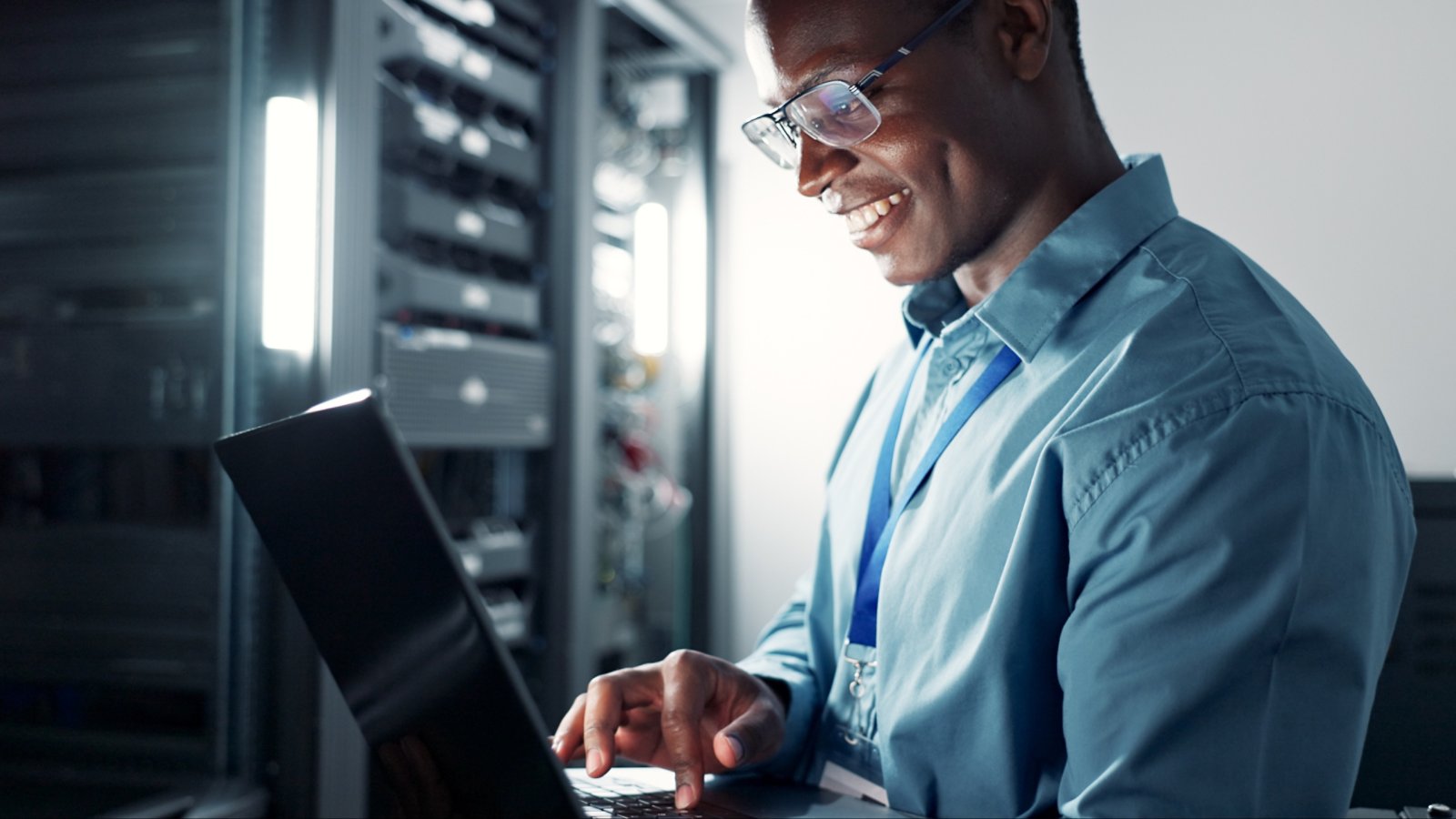Laptop, server room and black man on database network for maintenance, programming software or typing. Tech, data center and happy engineer on system for information, cybersecurity or cloud computing.