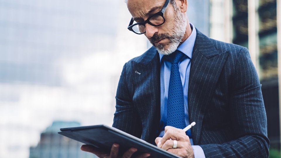 Handsome aged man in glasses and business suit using tablet with stylus while sitting against background of New York structures