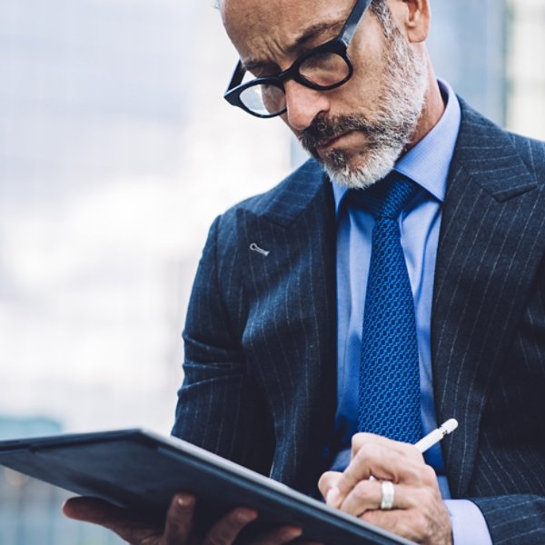 Handsome aged man in glasses and business suit using tablet with stylus while sitting against background of New York structures