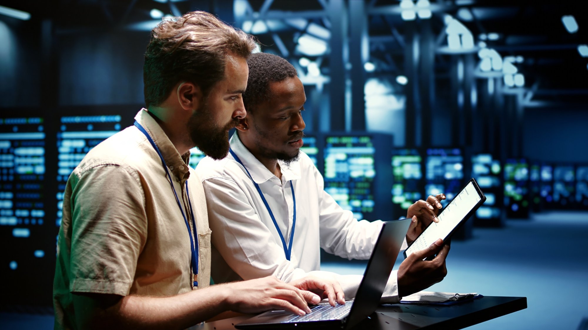 Coworkers running data center hardware diagnostic tests to determine and service software issues. Servicemen using laptop and tablet to fix critical systems, checking for blips