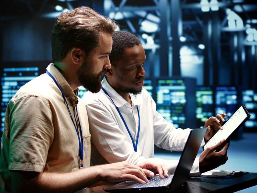 Coworkers running data center hardware diagnostic tests to determine and service software issues. Servicemen using laptop and tablet to fix critical systems, checking for blips