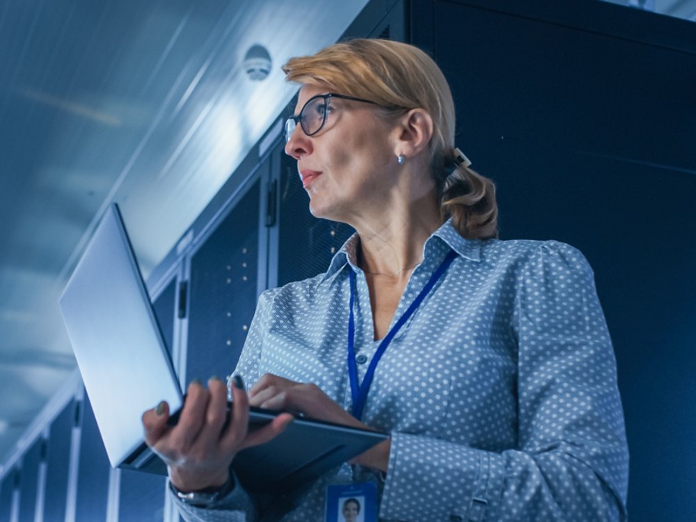 In Data Center: Low-Angle Portrait Shot of a Female IT Technician Running Maintenance Programme on Laptop, Controls Operational Server Rack. Modern High-Tech Telecommunications Operational Data Center