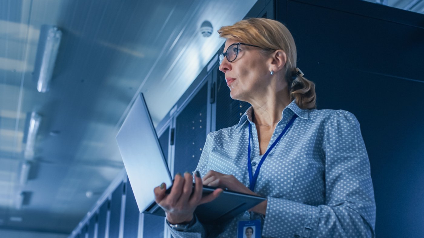 In Data Center: Low-Angle Portrait Shot of a Female IT Technician Running Maintenance Programme on Laptop, Controls Operational Server Rack. Modern High-Tech Telecommunications Operational Data Center