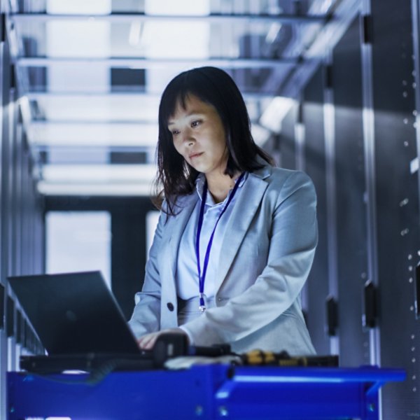 Asian Female IT Engineer Working on a Laptop on Tool Cart, She Scans Hard Drives.  She's in a Big Data Center Full of Rack Servers.