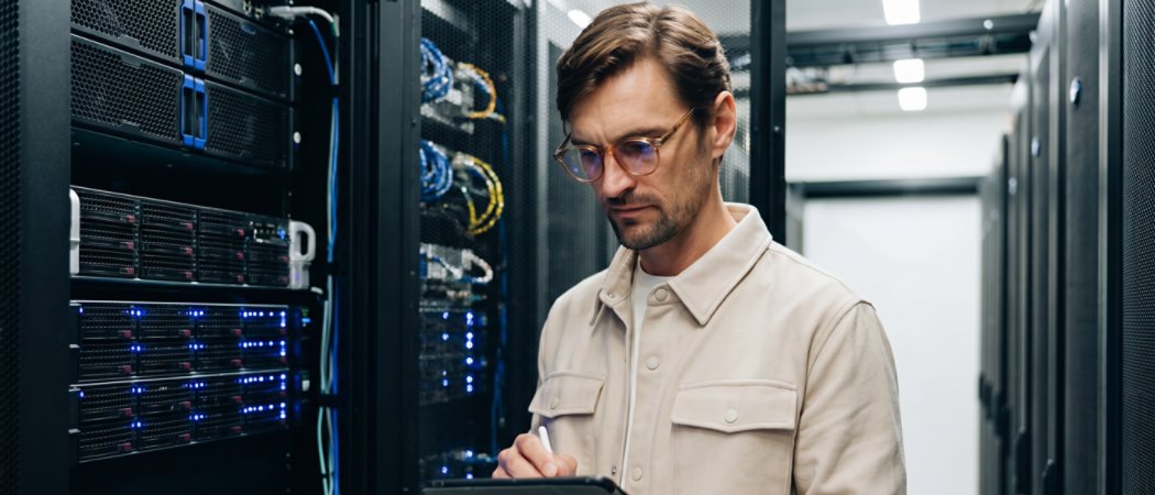 A person stands in a modern data center, holding a digital tablet while inspecting server racks. The environment is filled with black server cabinets, illuminated by blue indicator lights. The individual appears focused, suggesting a professional or technical activity. The setting is clean, organized, and features advanced technology infrastructure.