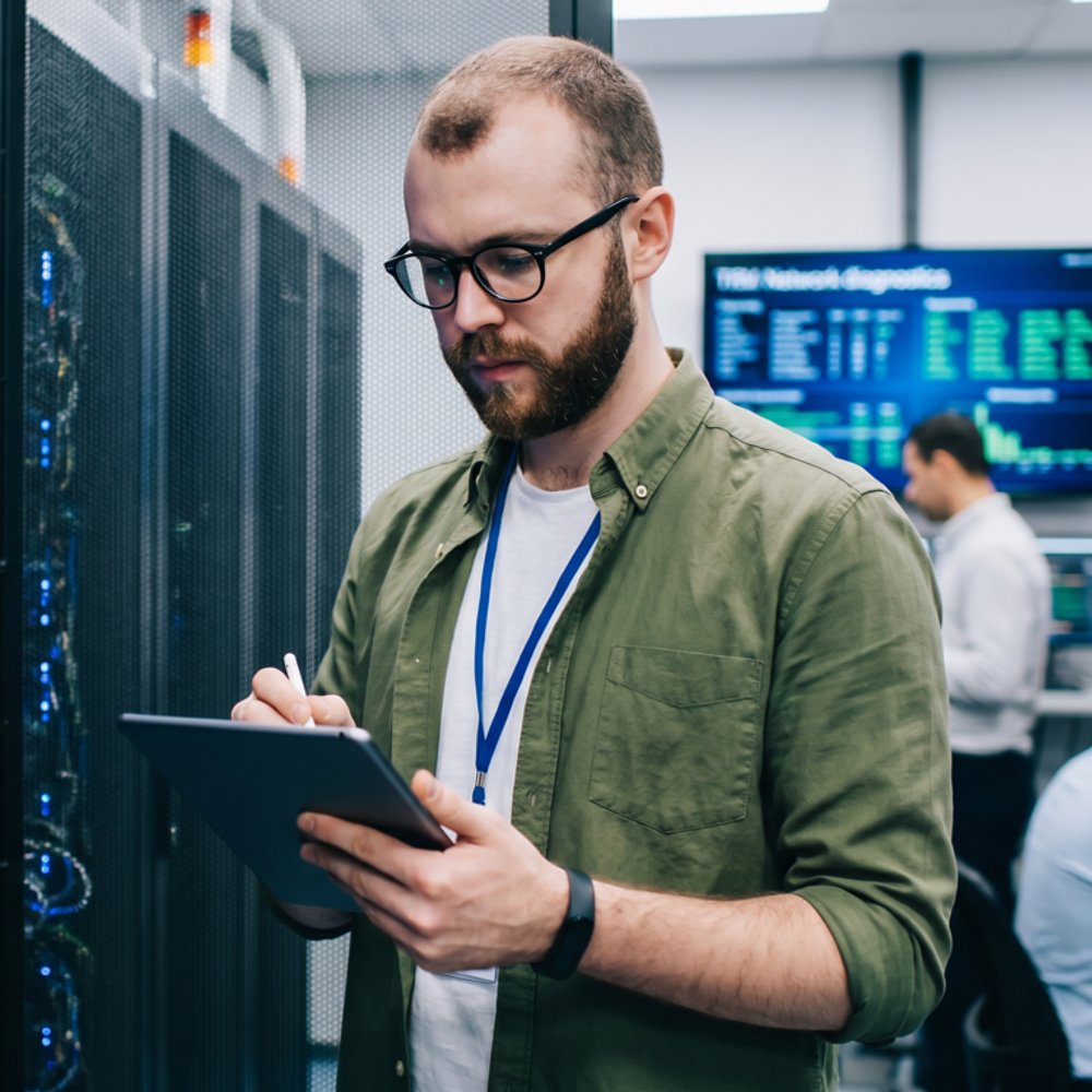 A person stands in a modern data center, holding a digital tablet and checking server racks. The environment is filled with advanced computer equipment and cables, with multiple screens displaying data in the background. Other individuals are working at desks, contributing to a busy, professional atmosphere. The setting is brightly lit, emphasizing a high-tech, efficient workspace.