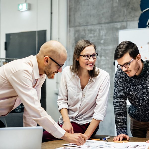 A team of four business people is standing in front a desk in a bright office room. They ambitiously point at documents while smiling and looking at the charts and notes.