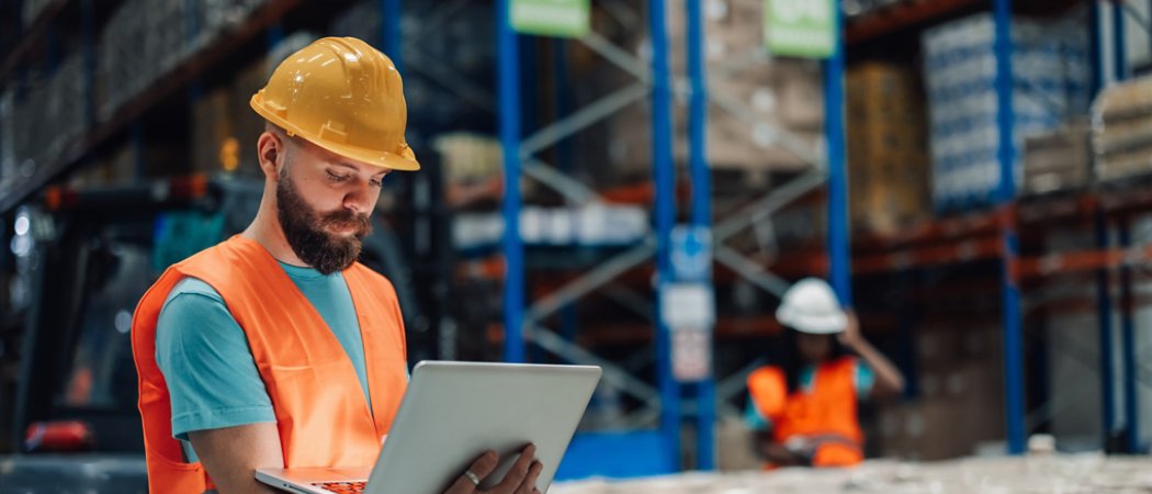 Warehouse worker wearing a safety vest and hardhat is holding a laptop, managing inventory and logistics in a large distribution center