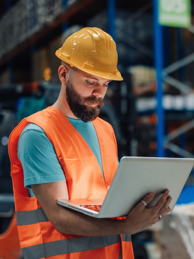 Warehouse worker wearing a safety vest and hardhat is holding a laptop, managing inventory and logistics in a large distribution center