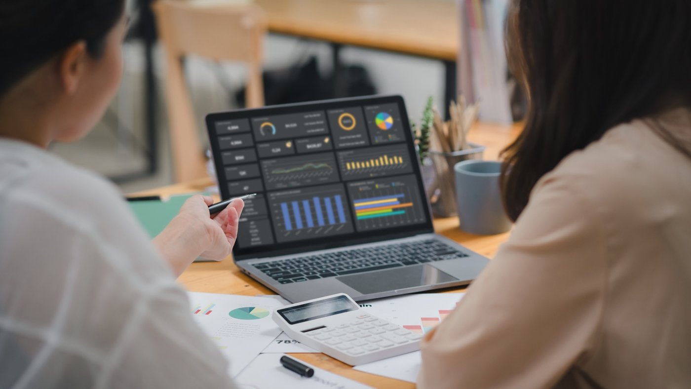 Two businesspeople or an accountant team are analyzing data charts, graphs, and a dashboard on a laptop screen in order to prepare a statistical report and discuss financial data in an office