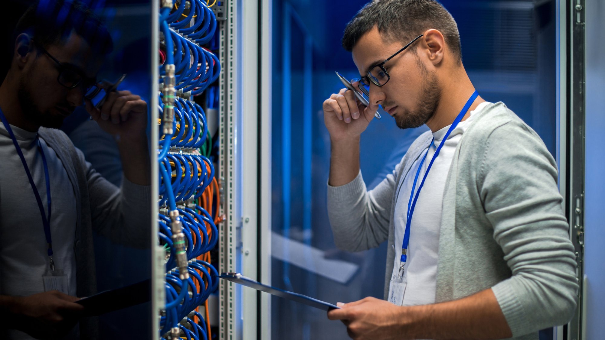 Side view  portrait of young man standing by server cabinet while working with supercomputer in data center and holding clipboard