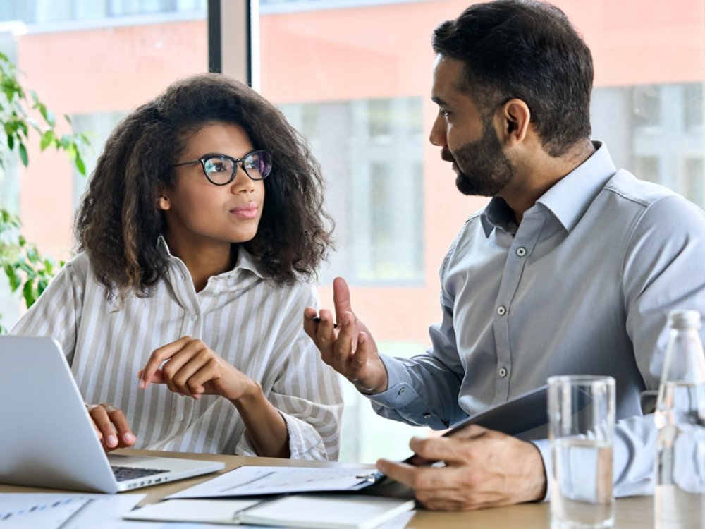 Businessman indian executive manager ceo talking to female African American coworker, using laptop. Diverse multicultural professional partners group discussing business plan at board room meeting.