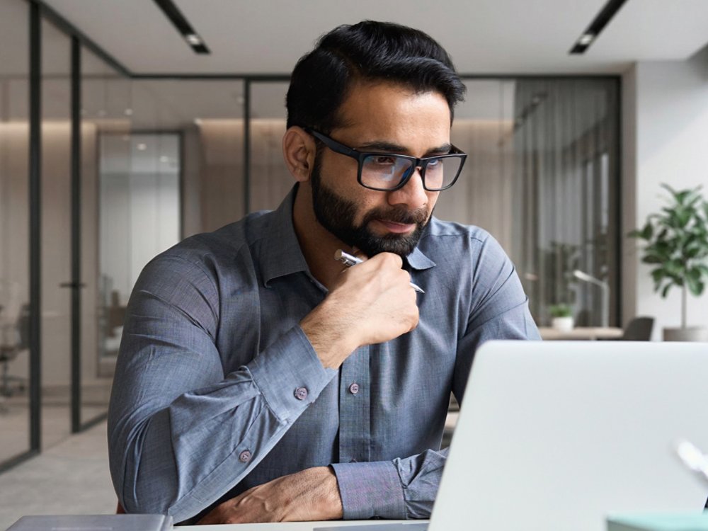 Young serious indian professional business man, focused ethnic male student wearing glasses working on laptop, remote studying using computer looking at screen watching seminar webinar at home office.