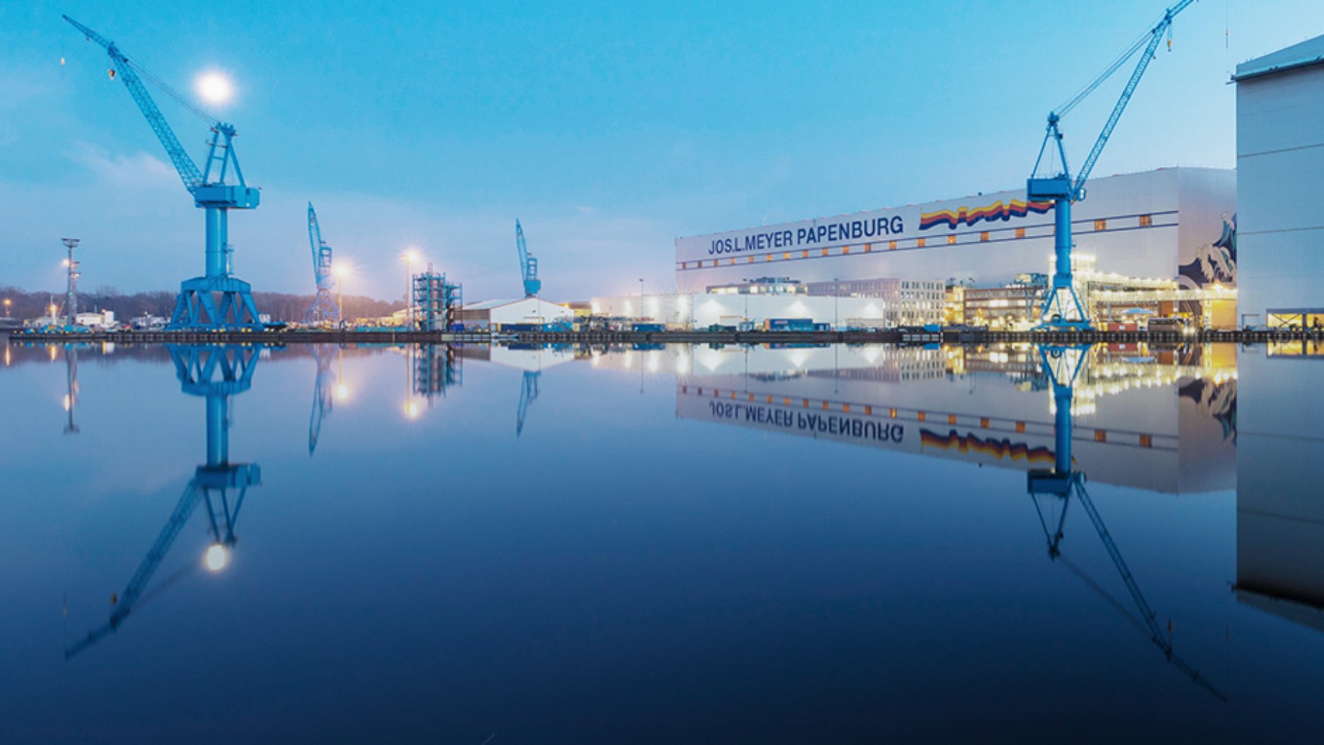 A large industrial shipyard stands beside a still body of water at dusk, with tall blue cranes dominating the skyline. The calm surface of the water creates a clear reflection of the cranes and buildings, enhancing the symmetry of the scene. Soft blue tones in the sky and water contrast with the warm lights from the shipyard structures. The text "JOSL MEYER PAPENBURG" is visible on a prominent building in the background.