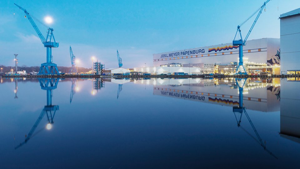 A large industrial shipyard stands beside a still body of water at dusk, with tall blue cranes dominating the skyline. The calm surface of the water creates a clear reflection of the cranes and buildings, enhancing the symmetry of the scene. Soft blue tones in the sky and water contrast with the warm lights from the shipyard structures. The text "JOSL MEYER PAPENBURG" is visible on a prominent building in the background.