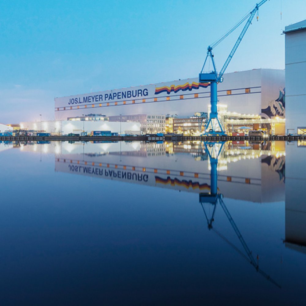A large industrial shipyard stands beside a still body of water at dusk, with tall blue cranes dominating the skyline. The calm surface of the water creates a clear reflection of the cranes and buildings, enhancing the symmetry of the scene. Soft blue tones in the sky and water contrast with the warm lights from the shipyard structures. The text "JOSL MEYER PAPENBURG" is visible on a prominent building in the background.
