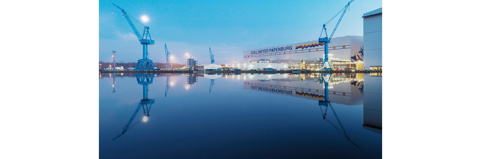 A large industrial shipyard stands beside a still body of water at dusk, with tall blue cranes dominating the skyline. The calm surface of the water creates a clear reflection of the cranes and buildings, enhancing the symmetry of the scene. Soft blue tones in the sky and water contrast with the warm lights from the shipyard structures. The text "JOSL MEYER PAPENBURG" is visible on a prominent building in the background.
