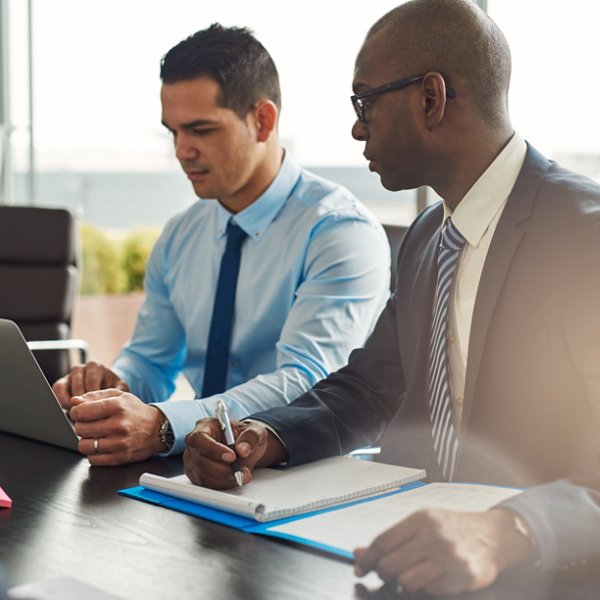 Two experienced business executives in a meeting seated at a table discussing paperwork and information on a laptop computer, one Hispanic, one African American