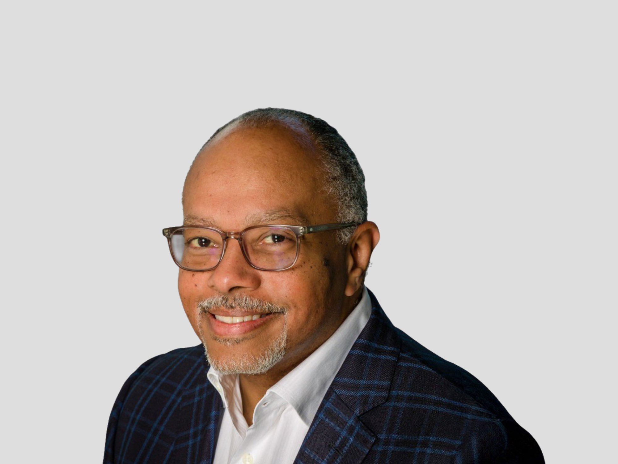 A person wearing a white shirt and a dark plaid blazer is shown against a plain gray background. The individual is also wearing glasses, adding a professional touch to the overall look. The setting is neutral and studio-like, emphasizing the formal attire.