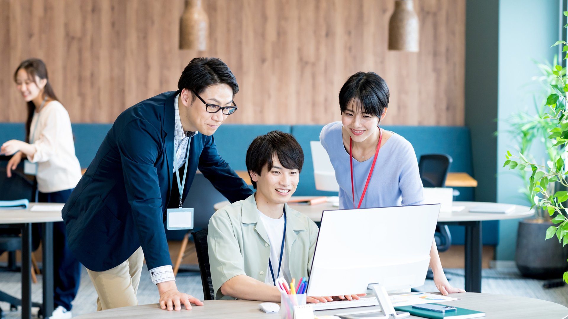 Three colleagues gather around a desktop computer, focusing on the screen and discussing work together. They are in a bright, contemporary open-plan office with wooden wall panels and pendant lights. Two people stand beside the seated coworker, all wearing ID badges on lanyards. Another person works in the background, adding to the professional, collaborative atmosphere.