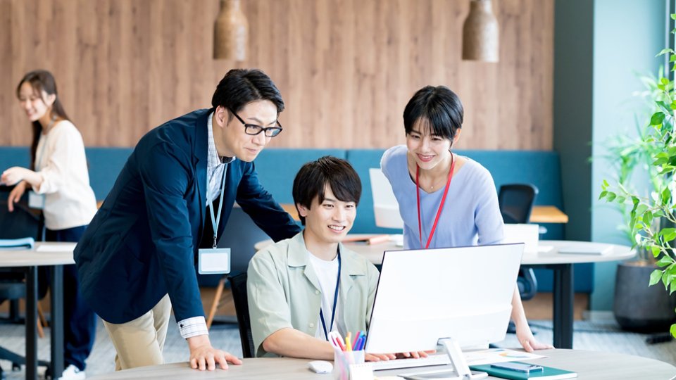Three colleagues gather around a desktop computer, focusing on the screen and discussing work together. They are in a bright, contemporary open-plan office with wooden wall panels and pendant lights. Two people stand beside the seated coworker, all wearing ID badges on lanyards. Another person works in the background, adding to the professional, collaborative atmosphere.
