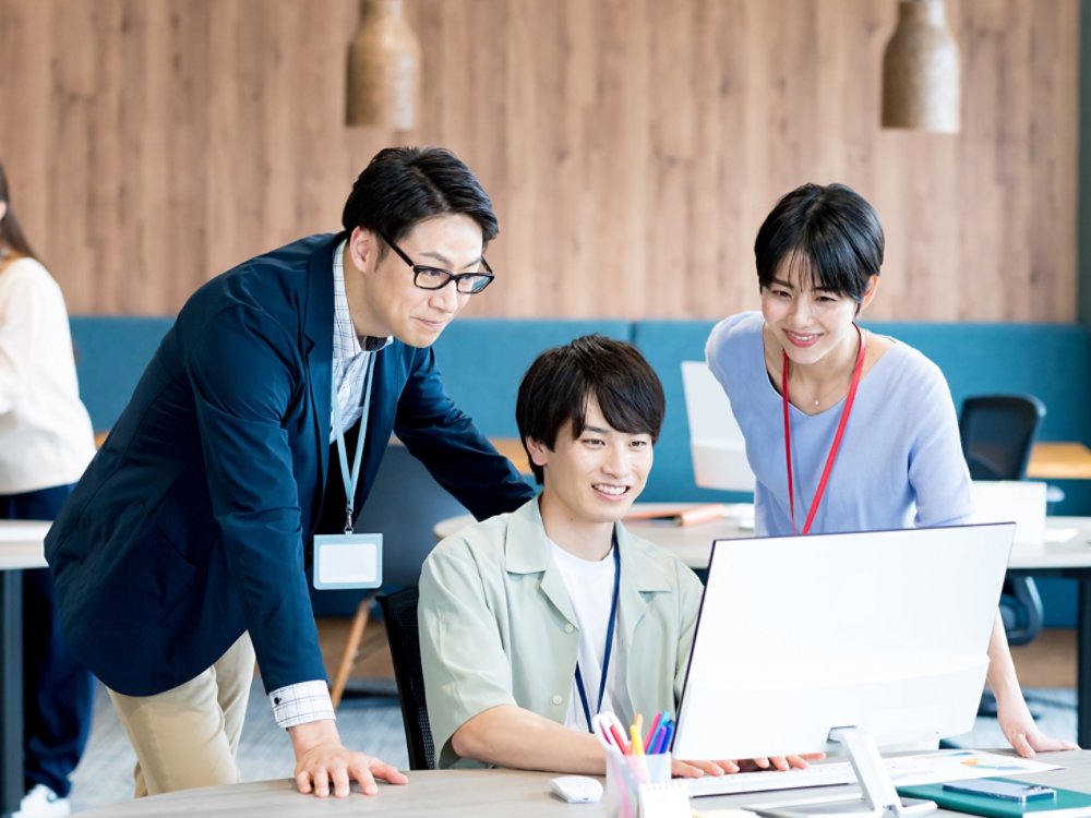 Three colleagues gather around a desktop computer, focusing on the screen and discussing work together. They are in a bright, contemporary open-plan office with wooden wall panels and pendant lights. Two people stand beside the seated coworker, all wearing ID badges on lanyards. Another person works in the background, adding to the professional, collaborative atmosphere.